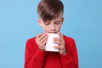 Cute boy drinking beverage from white ceramic mug on light blue background