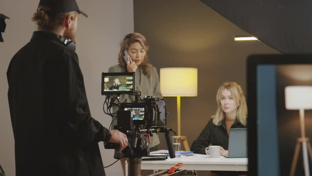 Medium shot of media production team getting ready for shooting in studio - videographer with camera on tripod discussing filming angle with model sitting at desk, and producer talking on mobile phone