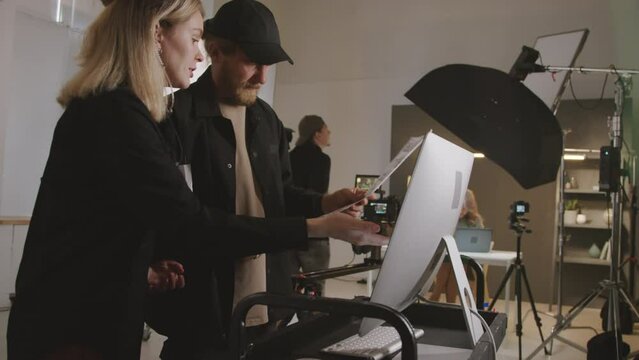 Medium shot of male stage director and female producer standing together in studio, viewing filmed footage on computer monitor, looking at storyboard and arguing, and videographer preparing camera