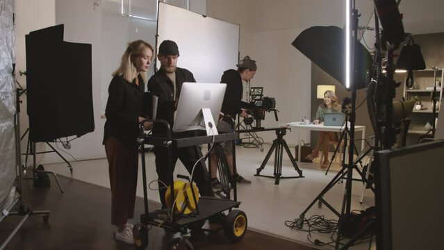 Full shot of media production crew filming in studio - videographer moving camera on dolly slider towards model, while director and technician are looking and discussing footage on computer monitor