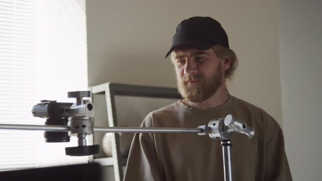 Medium shot of Caucasian male photographer tightening knob to adjust rack height and chatting to colleagues, while preparing to film flatlay in production studio