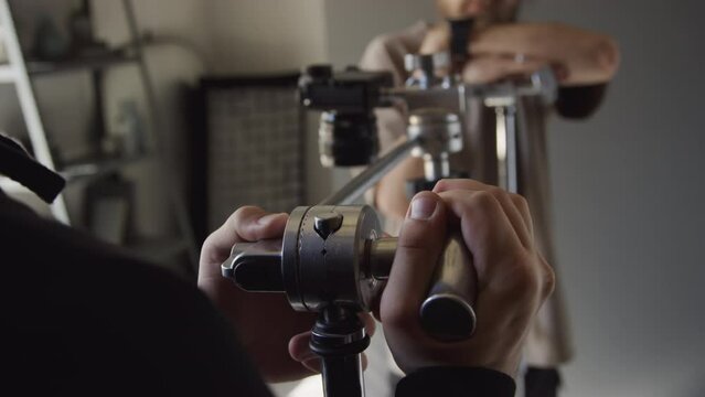 Medium shot of hands of anonymous male photographer tightening knob on horizontal camera rack and chatting to unrecognizable assistant, while preparing to shoot flatlay in studio