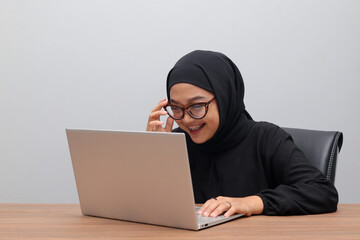 Portrait of attractive Asian hijab woman working on her laptop in office. Muslim girl adjusting glasses to see the screen clearly. Employee and freelance worker concept.