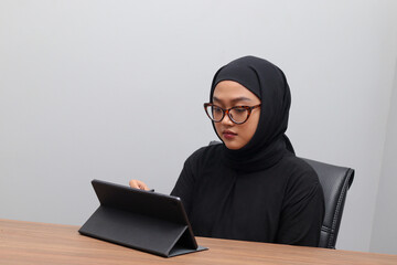 Portrait of attractive Asian hijab woman working on her tablet. Muslim girl doing task in office. Employee and freelance worker concept.
