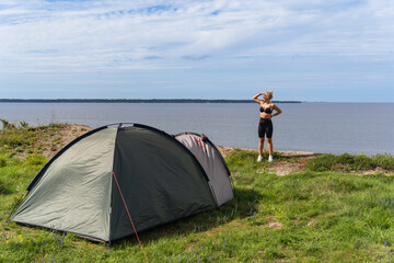 A beautiful slender girl stands near a tent on a steep seashore in summer.