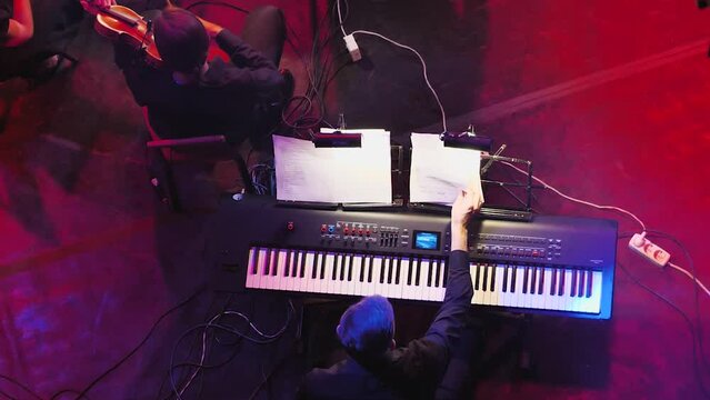A man at a synthesiser altering notes while violinists play nearby. Keyboard player in the symphony orchestra. View from above. Light accompaniment to the show