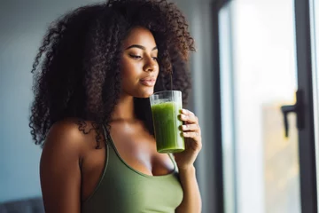 Poster Young attractive african american woman drinking her detox juice after workout, beautiful athletic female drinking green juice at home © VisualProduction