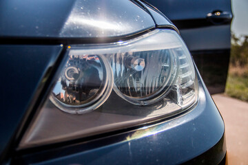 The double headlight of a dark car glows and the sun reflects on the hood close-up