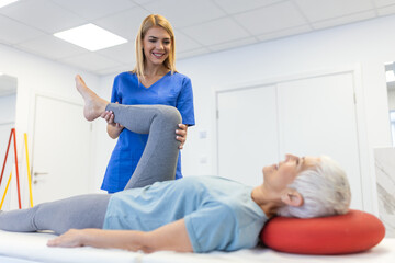 close up of female physiotherapist is helping senior elder woman stretching her hamstring and doing thigh or leg rehabilitation in exercise room - she is lying on massage bed - obrazy, fototapety, plakaty