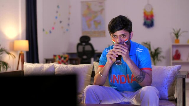 An Indian man in a blue cricket jersey watching a live cricket match on TV or television - cricket fever  World Cup series. Indian cricket fan wearing an Indian jersey - excited  cheering for a tea...