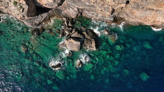 Top down drone shot of crystal clear water with rocks next to stone cliff and birds flying by