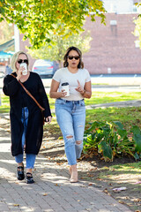 Two women in their 30s walking outside talking, drinking coffee