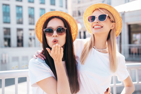 Two young beautiful smiling hipster female in trendy summer white t-shirt and jeans clothes. Carefree women posing in the street. Positive models having fun. Cheerful and happy. In hat. give air kiss
