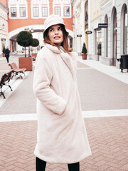Obraz premium Young beautiful smiling lady wearing trendy white faux fur coat. Stylish woman posing in the street in winter. Cheerful and happy model in panama. Cold autumn weather. Shopping mall