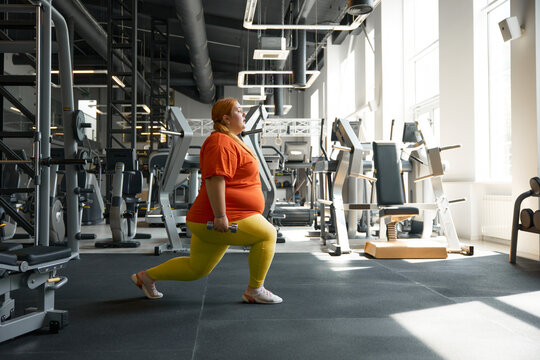 Confident obese woman doing forward lunges with dumbbells at gym