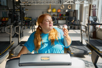 Front view on funny obese woman eating cake while training on treadmill at gym