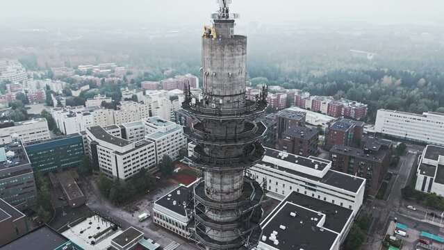 Aerial shot of a bleak industrial concrete television and radio link tower in Pasila, Helsinki, Finland on a bright and foggy day. Camera orbiting around, apartment buildings and forest in background.