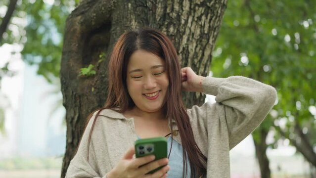 Happy asian young woman raising hand for celebrating moment while using smartphone standing under the tree at public park. Female reading a good news on smartphone raising hand for celebrating