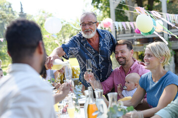 Grandfather opening bottle of champagne, pouring a champagne into glasses. Senior man making celebratory toast at outdoor summer garden party.