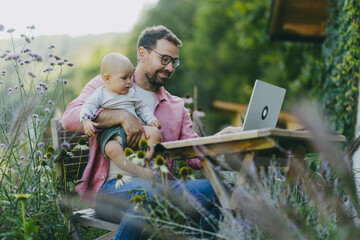 Father holding baby while working on laptop in garden. Businessman working remotely from outdoor home office and taking care of little son. Life work balance with kid.