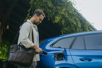 Portrait of a man charging electric car in front of his house, plugging the charger into the charging port. The man is unplugging charger from the fully charged car before going to the office.