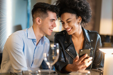 A young man and woman, seated at a chic restaurant, share a joyful moment looking at a smartphone....