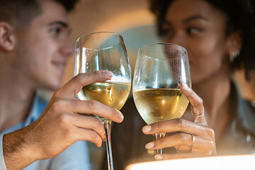 An intimate close-up of a young man and woman toasting with glasses of white wine. Their focused...