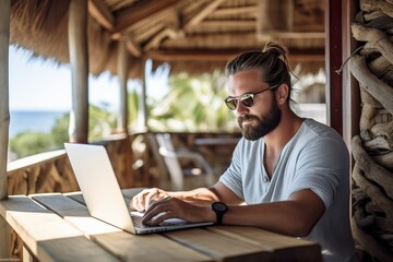 Man working remotely on his computer in a wooden cabin in a tropical location - obrazy, fototapety, plakaty