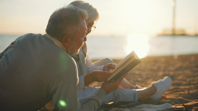 Senior gray haired man woman reading book together sitting on sandy sea beach at sunset in evening. Happy marriage, old family spending enjoy time. Love couple relationship, leisure, resting concept.