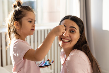 Smiling mother and preschool daughter doing makeup together, excited little girl, holding makeup brush, applies powder to mother's face, happy child applying blush, having fun playing with mom at home