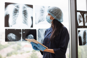 Focused female medical worker in face mask and scrub top posing with digital tablet near x-ray scans fixed on window glass. Qualified hindu surgeon in medical gloves studying injuries.