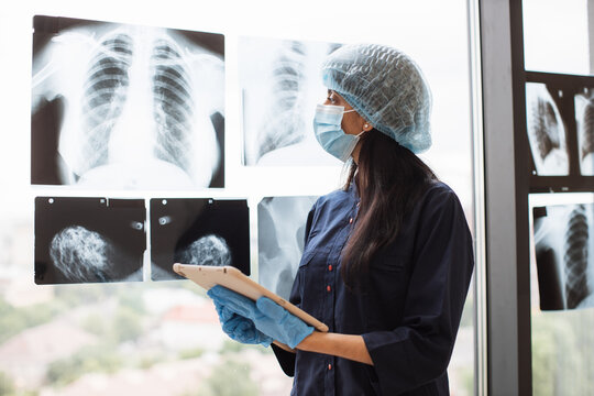 Focused female medical worker in face mask and scrub top posing with digital tablet near x-ray scans fixed on window glass. Qualified hindu surgeon in medical gloves studying injuries.