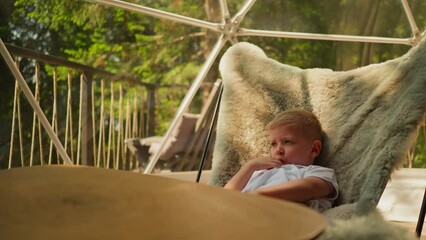 Thoughtful little boy sits in fur chair and watches nature surrounding glamping site. Curious child explores forest while sucking thumb and basking in sun