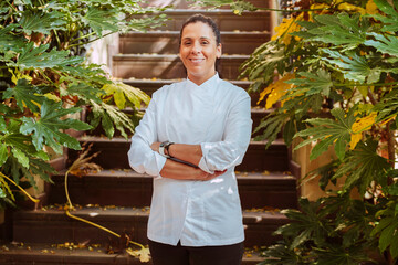 Female chef wears white coat, broadly smiles posing with arms crossed outdoors in front of stairs and plants with big green leafs. Restaurant worker, culinary gourmet, pastry chef.