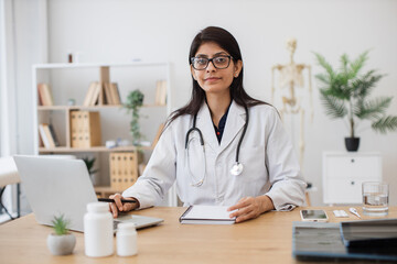 Female general practitioner in medical gown sitting and keeping hand on notebook while working with wireless laptop. Indian woman performing task professionally in field of medicine at office.