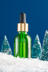 Winter season skin care concept. Closeup of green dropper bottle with face serum or oil and Christmas trees with snow on blue background with blank space. Unbranded mockup for adv