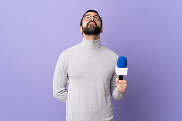 Adult reporter man with beard holding a microphone over isolated purple background and looking up