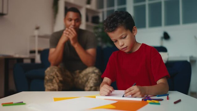 Man in military uniform looking at little son drawing picture, family time