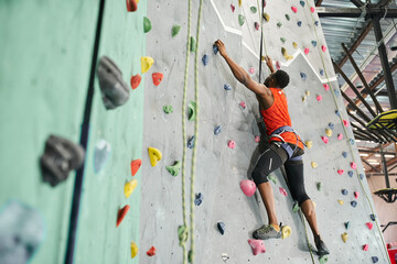 handsome african american man in orange shirt gripping on boulders and looking up, sportsman