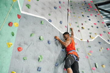 sporty young african american man using safety rope and harness to climb up rock wall, bouldering