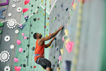 young african american man climbing up bouldering wall with safety rope and alpine harness