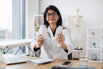 Friendly female therapist in glasses showing white jar of medicine while sitting at desk. Experienced indian woman in white coat recommending best alternative of treating at doctor's office.