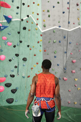 vertical shot of sporty african american man in orange shirt with alpine harness near rock wall