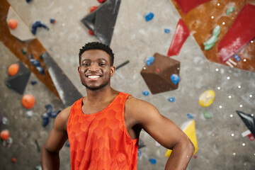 handsome jolly african american man in orange shirt standing next to rock wall and smiling at camera - obrazy, fototapety, plakaty