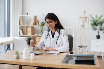 Competent doctor using modern laptop and gesturing during online video consultation. Indian woman in medical uniform telling about home treatment plan to sick patient while sitting at table.