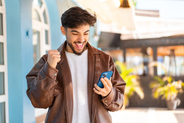 Handsome Arab man at outdoors using mobile phone and doing victory gesture