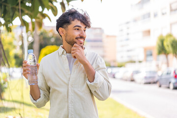 Young Arabian handsome man with a bottle of water at outdoors thinking an idea and looking side