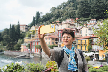 Happy old woman taking selfie with a smart phone in Varenna, Italy. Lake Como city - Older woman enjoying summer holidays - Holidays, mature people and travel concept.