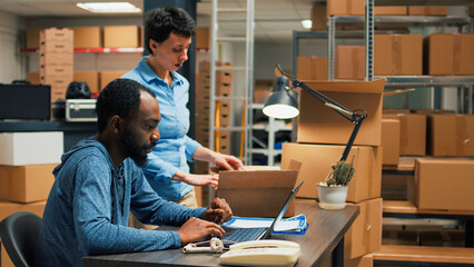 Young partners checking quality of merchandise packed in cardboard boxes, preparing order to ship to customers. Team of employees working on shipping products from warehouse shelves. - obrazy, fototapety, plakaty
