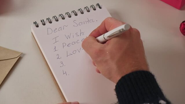 Male Person Writing Letter for Santa Claus, hand holding pen. Dear Santa. Man in Christmas Sweater making Wish List. Xmas Wishing. He wants Love, Peace, Health, Money. Desktop with Holiday decoration.